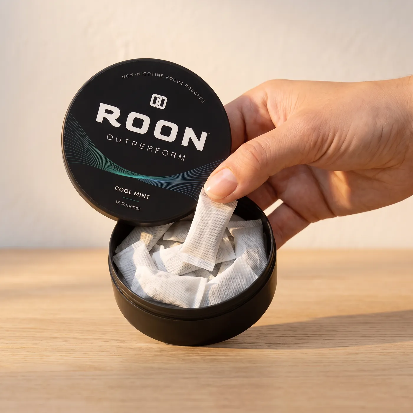 A hand lifting one flat white Roon Cool Mint pouch from an open matte black tin, on a clean cream background.