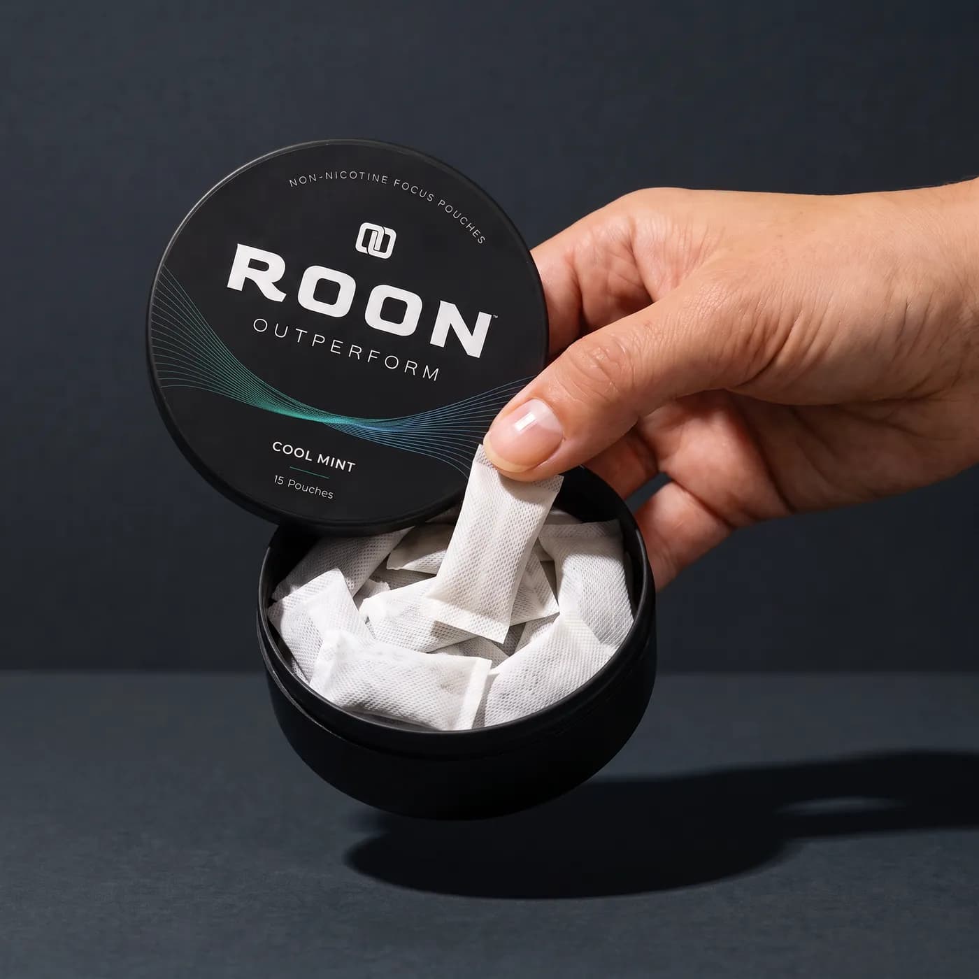 A hand lifting one flat white Roon Cool Mint pouch from an open matte black tin, on a clean cream background.