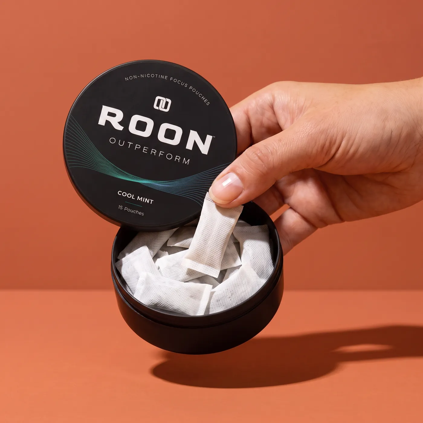 A hand lifting one flat white Roon Cool Mint pouch from an open matte black tin, on a clean cream background.