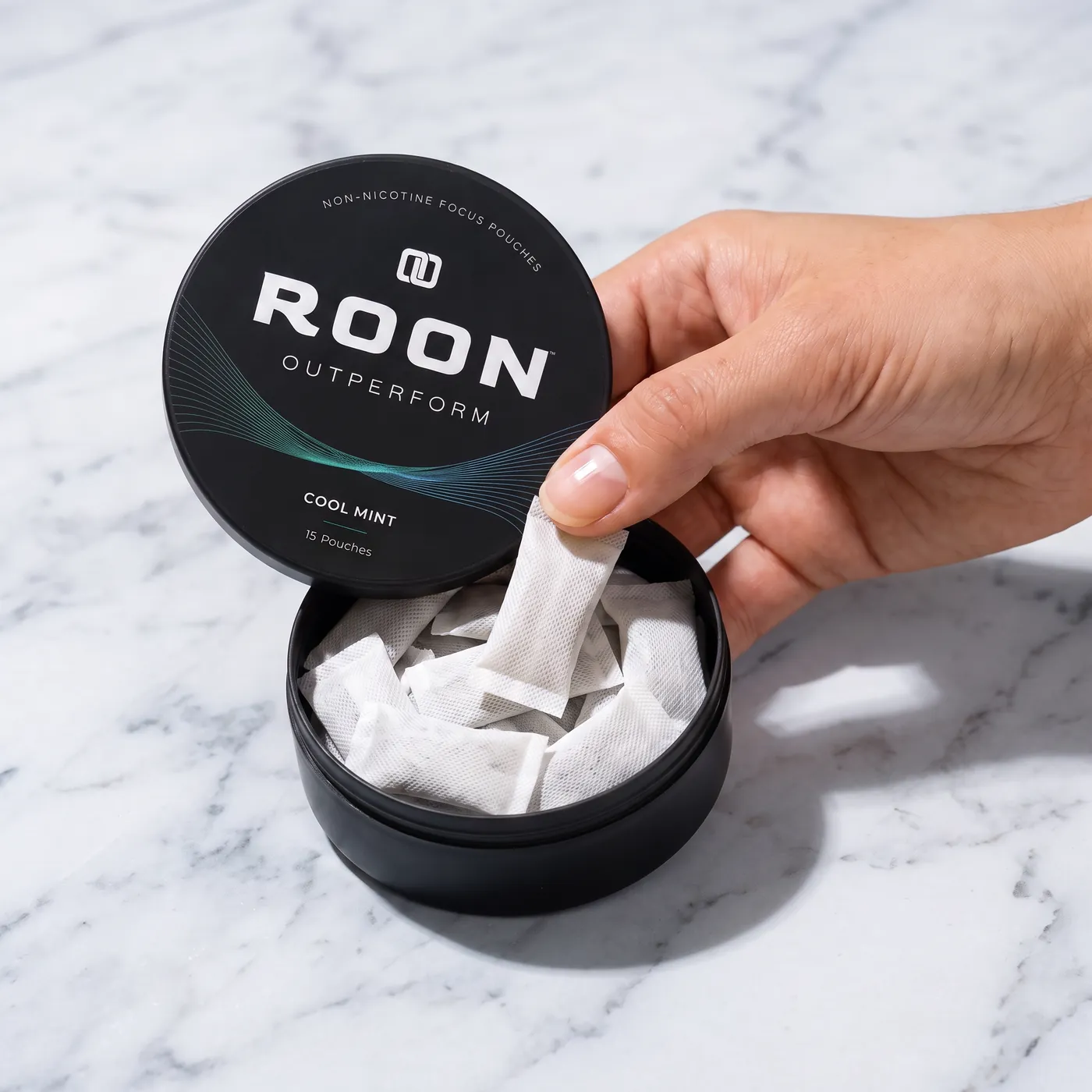 A hand lifting one flat white Roon Cool Mint pouch from an open matte black tin, on a clean cream background.