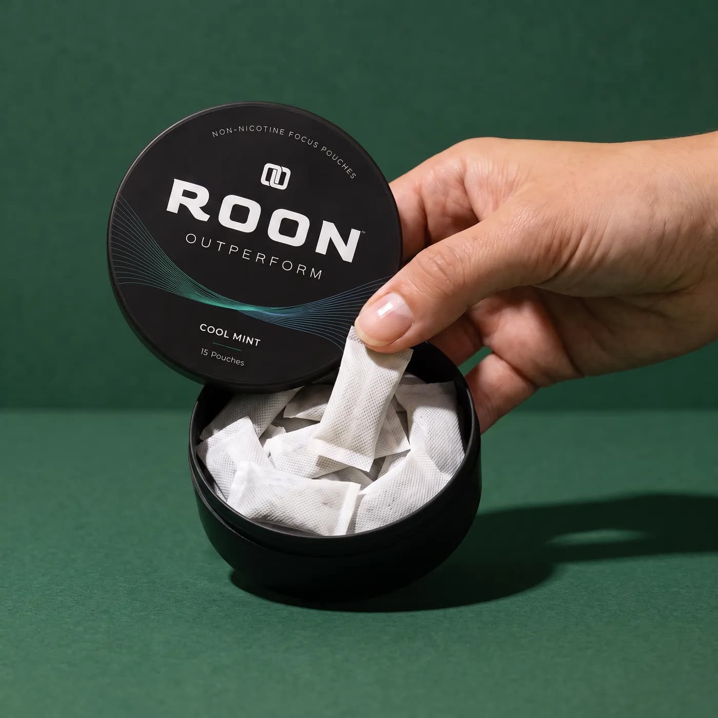 A hand lifting one flat white Roon Cool Mint pouch from an open matte black tin, on a clean cream background.