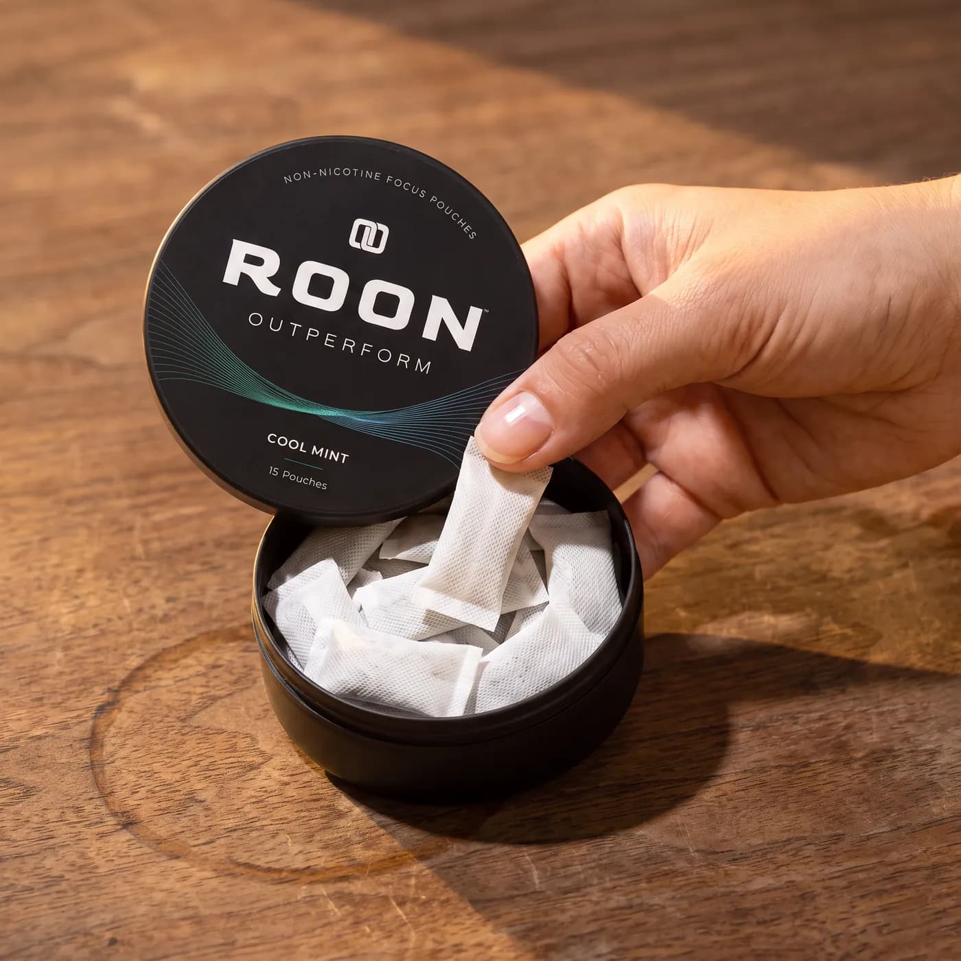 A hand lifting one flat white Roon Cool Mint pouch from an open matte black tin, on a clean cream background.