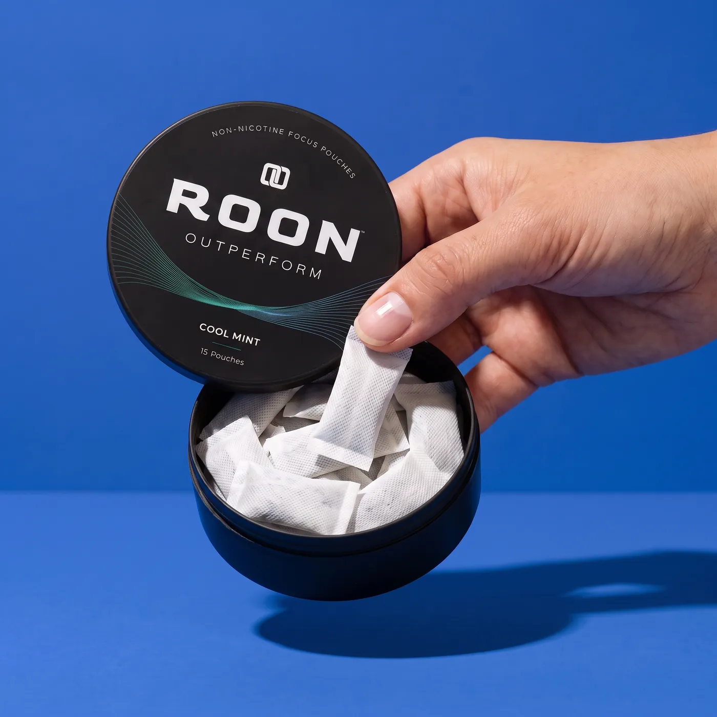 A hand lifting one flat white Roon Cool Mint pouch from an open matte black tin, on a clean cream background.