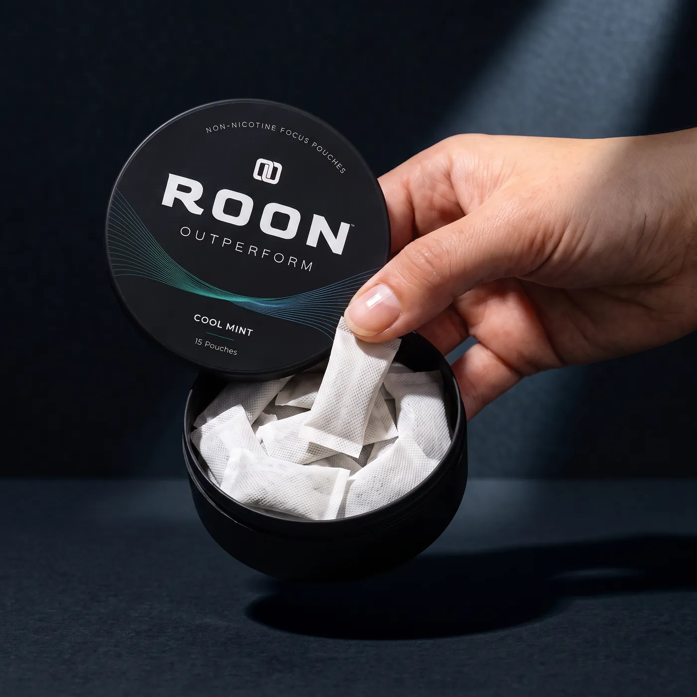 A hand lifting one flat white Roon Cool Mint pouch from an open matte black tin, on a clean cream background.