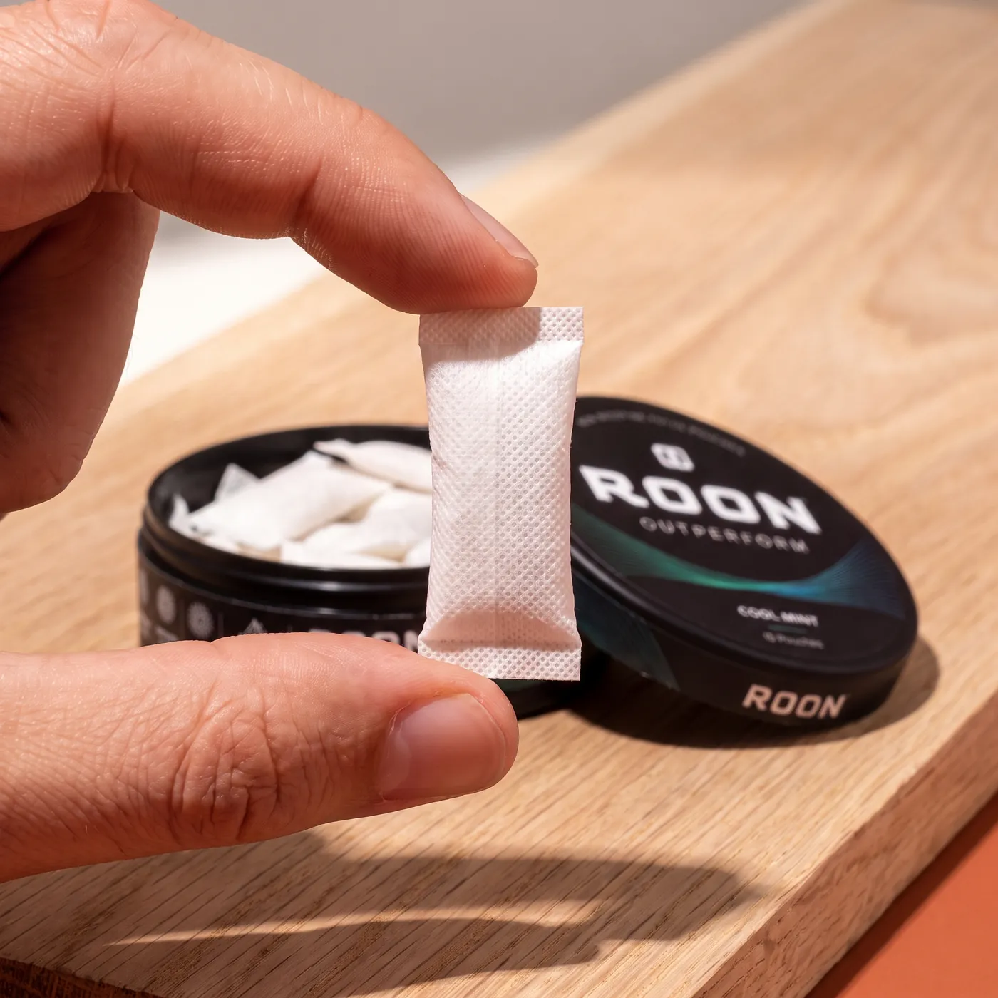 Close view of a single flat white Roon caffeine pouch held between two fingertips, showing the woven fabric.