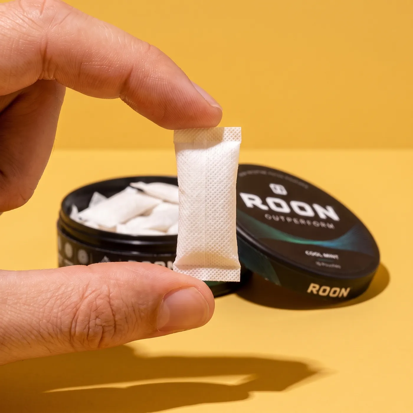 Close view of a single flat white Roon caffeine pouch held between two fingertips, showing the woven fabric.