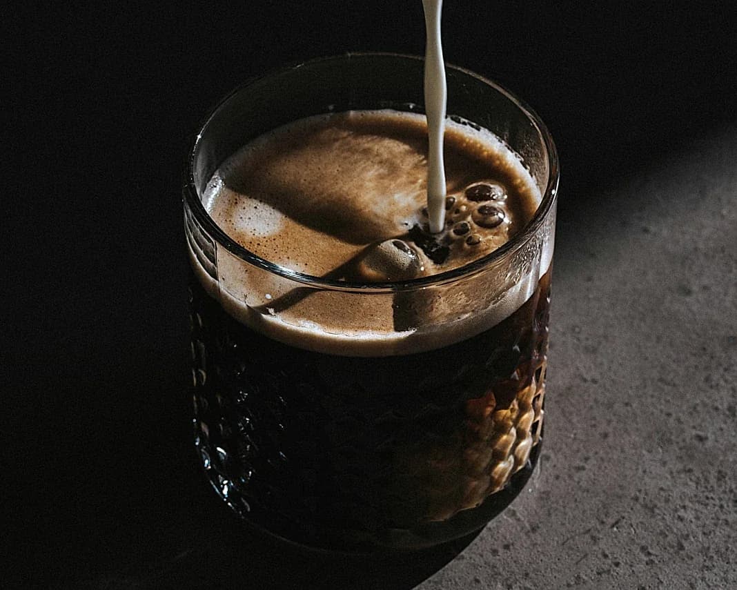 Milk being poured into a glass of iced coffee on a dark countertop